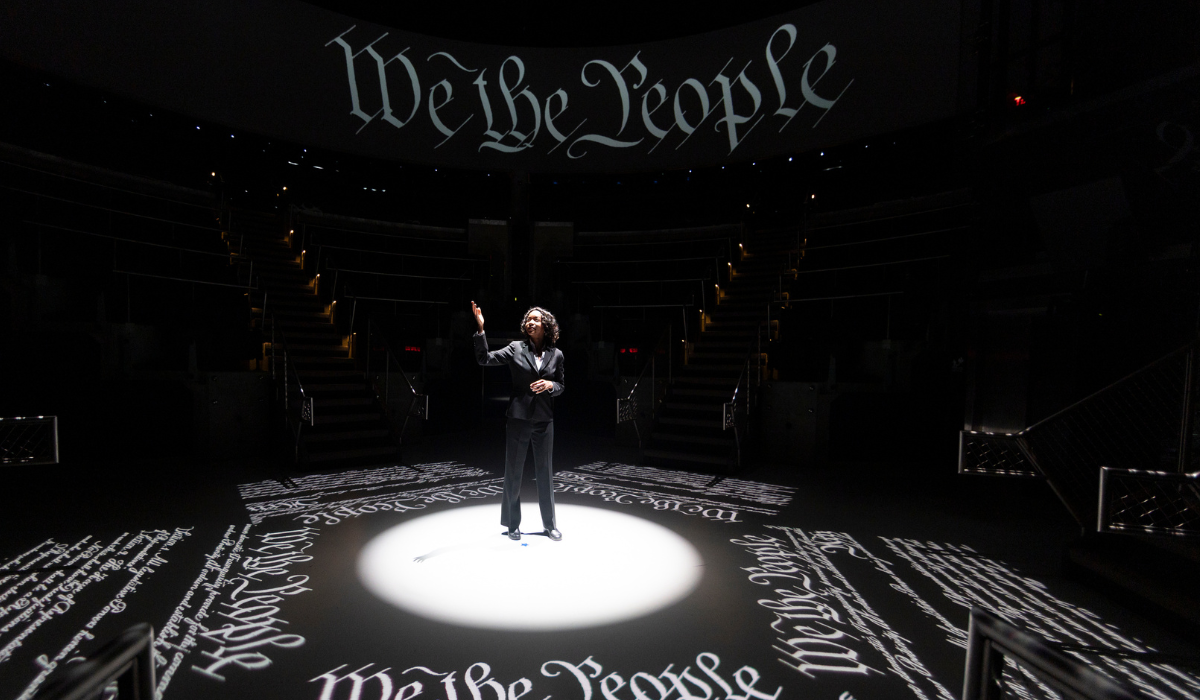 A speaker stands in a spotlight on a dark stage, surrounded by projections of the phrase 