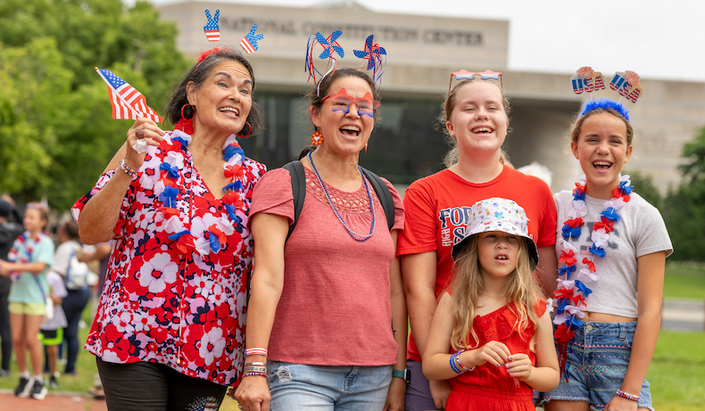 A group of five people wearing patriotic red, white, and blue accessories smile in front of the National Constitution Center.