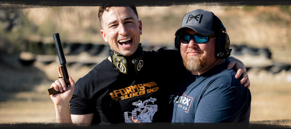 Two people wearing hearing protection smile together at an outdoor shooting range, with one holding a suppressed handgun.