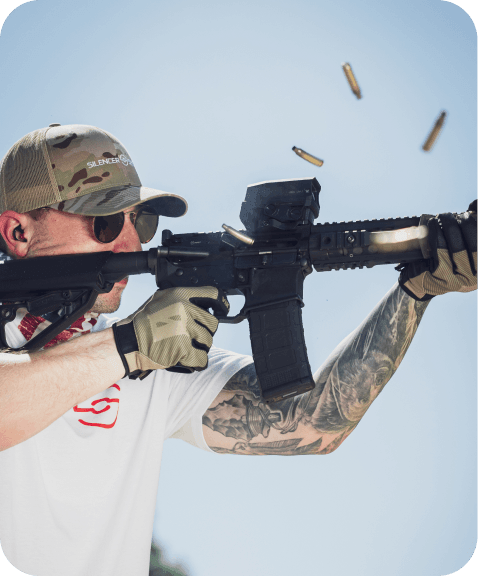 A shooter in protective gear fires a tactical rifle outdoors, with spent shell casings visible in the air.