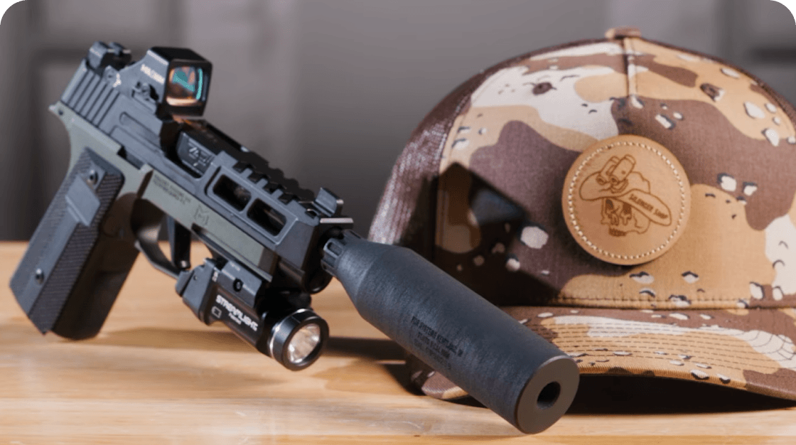 Black handgun with suppressor, mounted flashlight, and red dot sight placed on wooden surface beside camouflage cap.