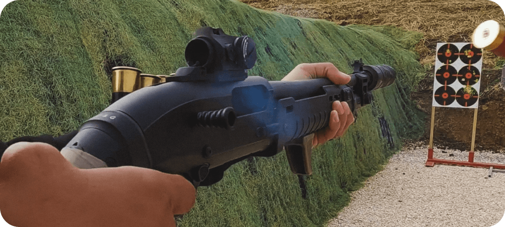 Shooter aiming a suppressed shotgun with a red-dot sight at a paper target on an outdoor range