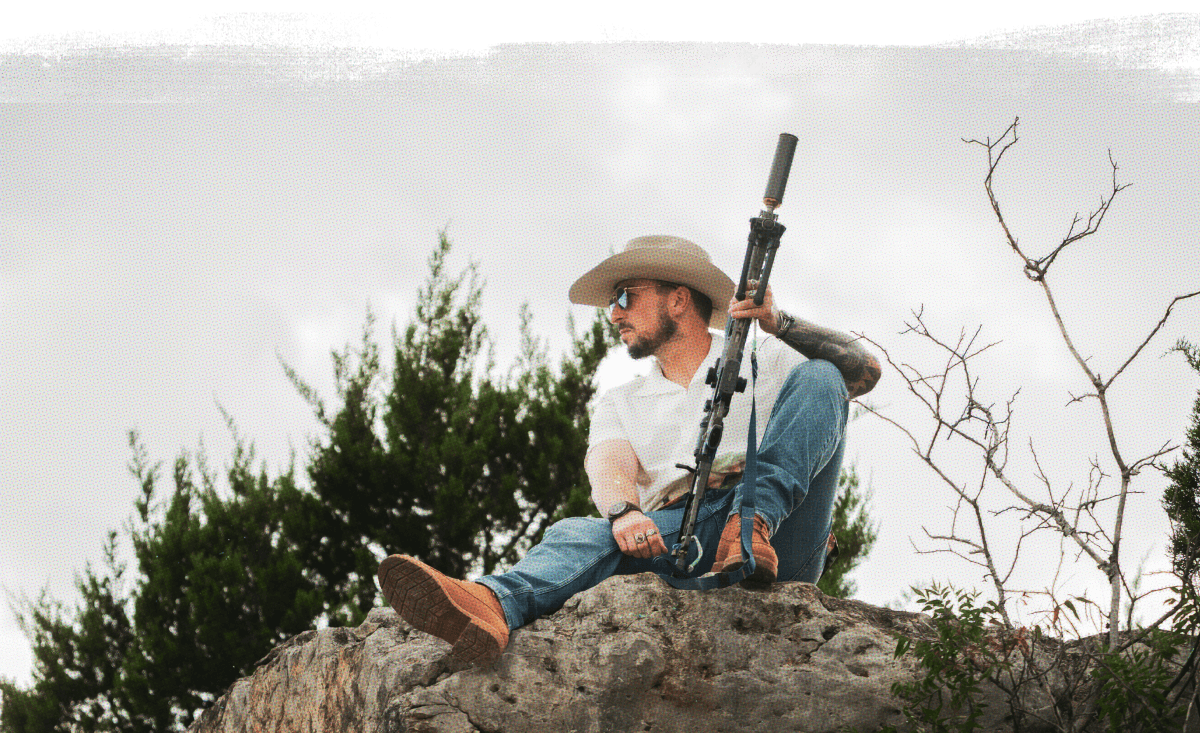 Individual in hat and sunglasses holding rifle with suppressor on rocky terrain.