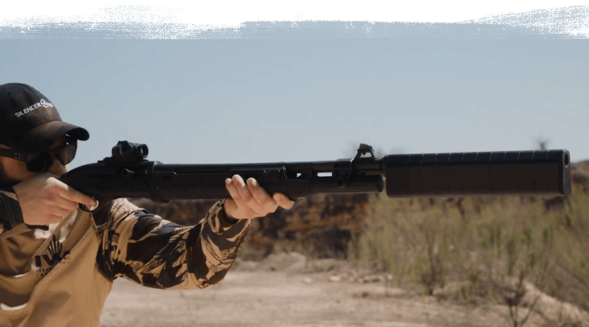Hunter in camo aiming a suppressed shotgun with a red-dot sight in a desert landscape