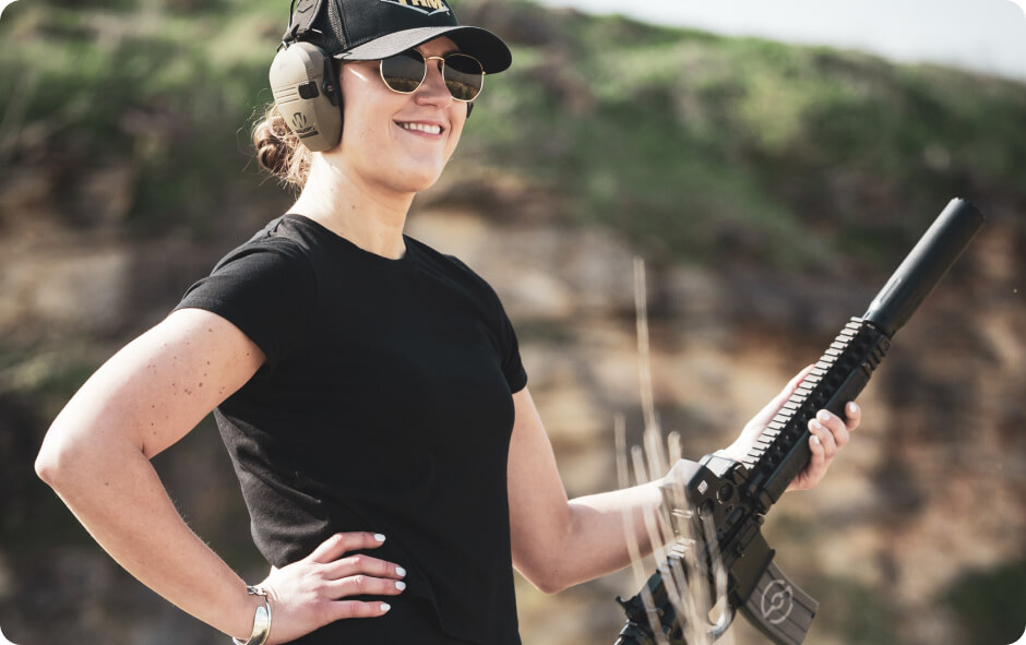 Individual in gloves and cap firing tan rifle with scope, laser module, suppressor, and red patterned magazine, muzzle flash visible at outdoor range.
