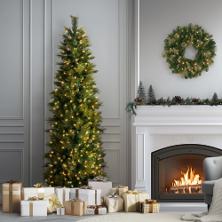 Slim Christmas tree with warm lights beside a white fireplace decorated with garland and a wreath, surrounded by gold and white presents.