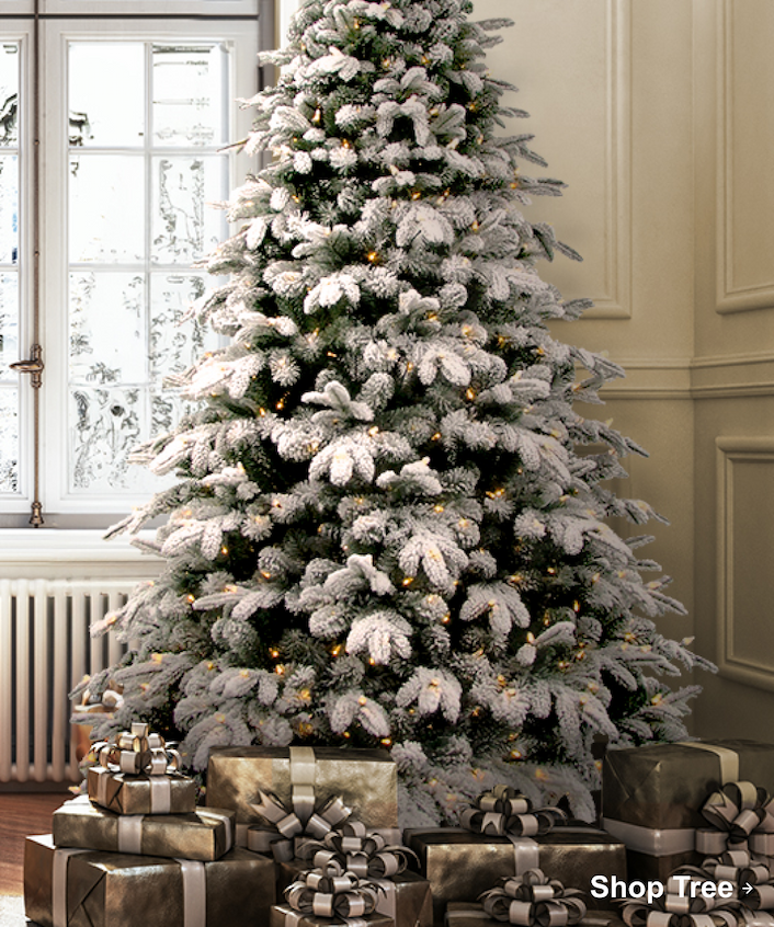Flocked Christmas tree with warm lights surrounded by silver and gold wrapped presents in a bright room.