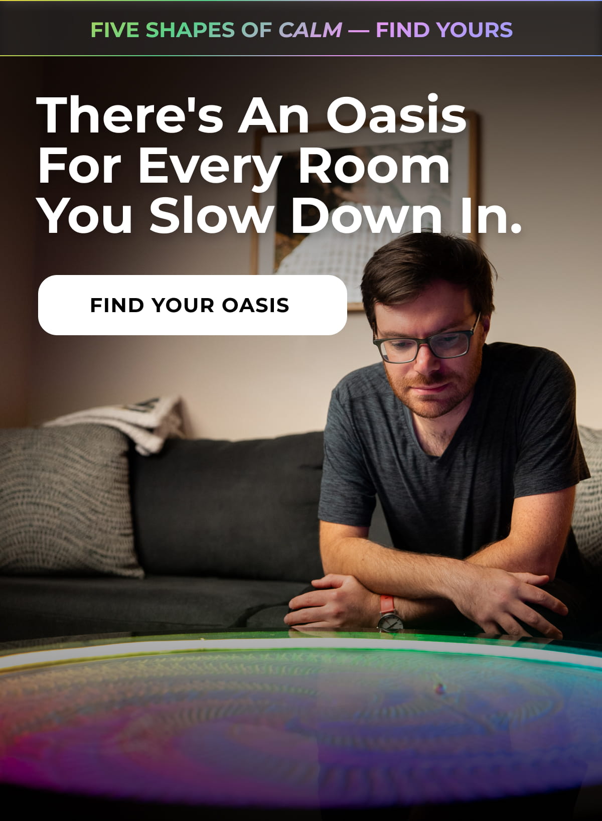 Man relaxing on a sofa while watching a colorful illuminated Oasis kinetic sand table.