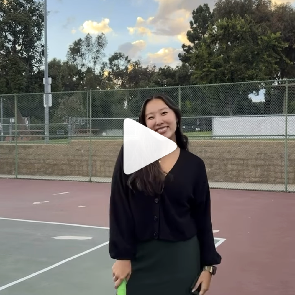 [A young woman with dark hair stands, smiling, on a tennis court, holding a tennis racket by her side. A white triangle-shaped "play" icon is over the image.]