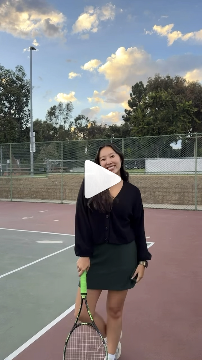 [A young woman with dark hair stands, smiling, on a tennis court, holding a tennis racket by her side. A white triangle-shaped "play" icon is over the image.]
