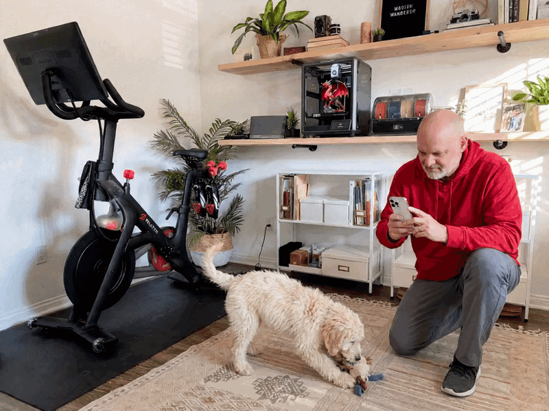 A man in a red hoodie checking his phone next to his Peloton bike and 3D printer while his dog plays on the floor.