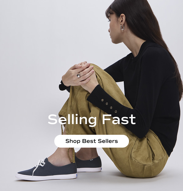 A woman sitting with her knees bent wearing a black top with buttons at the cuffs, cargo pants, and the Keds Champion Point Low Canvas in navy. The email says Selling Fast. Shop Best Sellers.