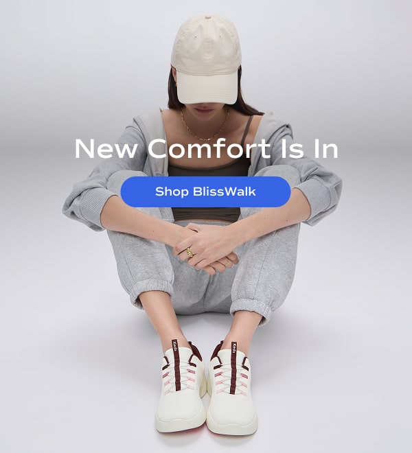 A woman in a beige ball cap, matching grey sweatshirt and sweatpants, and the BlissWalk Slip-On in snow white knit with pink and mauve accents. New Comfort Is In. Shop BlissWalk.