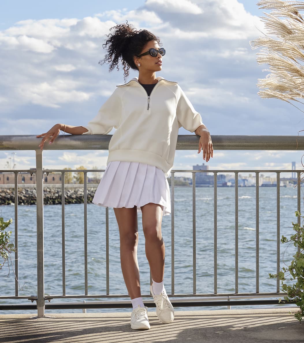 A woman leaning against a metal railing with a seascape behind her. She is wearing sunglasses, a cream half-zip sweater, white pleated skirt, white socks, and the Keds BlissWalk Jersey Lace-Up in cream.