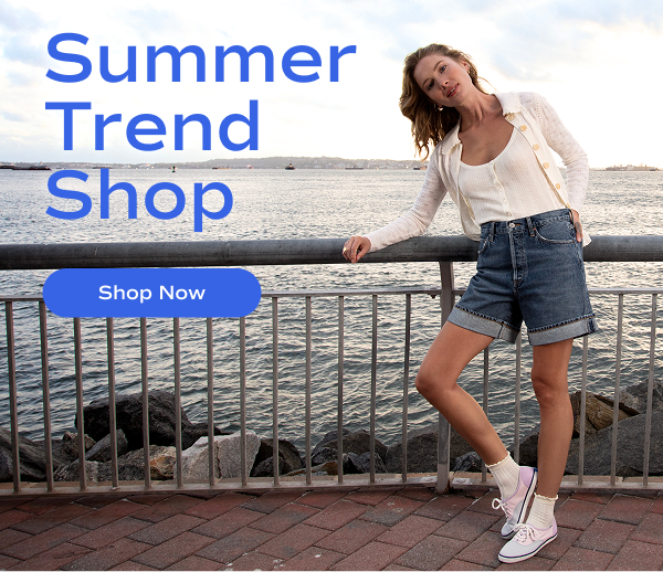 A woman leaning against a metal fence with a seascape behind her, she is wearing an open white cardigan with matching tank top, denim cuffed shorts, ankle socks and the Keds Champion Canvas in color-block pastel. Text on the image to the left of the woman reads, "Summer Trend Shop" along with a "Shop Now" button.