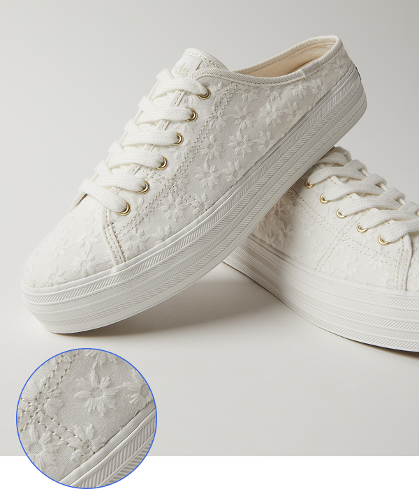 A close-up view of two Keds Triple Kick Starburst Eyelet mules in snow white. The shoes feature an all-over white daisy eyelet embroidered fabric upper, white cotton laces threaded through gold-toned eyelets, and a white platform sole. A circular inset in the lower left corner provides a detailed view of the floral-embroidered fabric and stitching on the side of the shoe and the textured rubber of the sole.