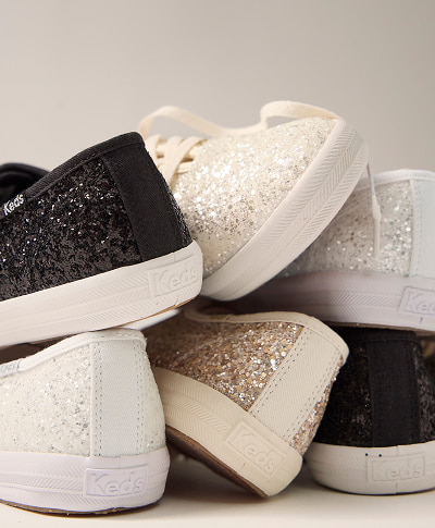 A zoomed-in view of a pile of Keds Champion Glitter Lace-Ups in a variety of shades including black, white, cream, and gold.