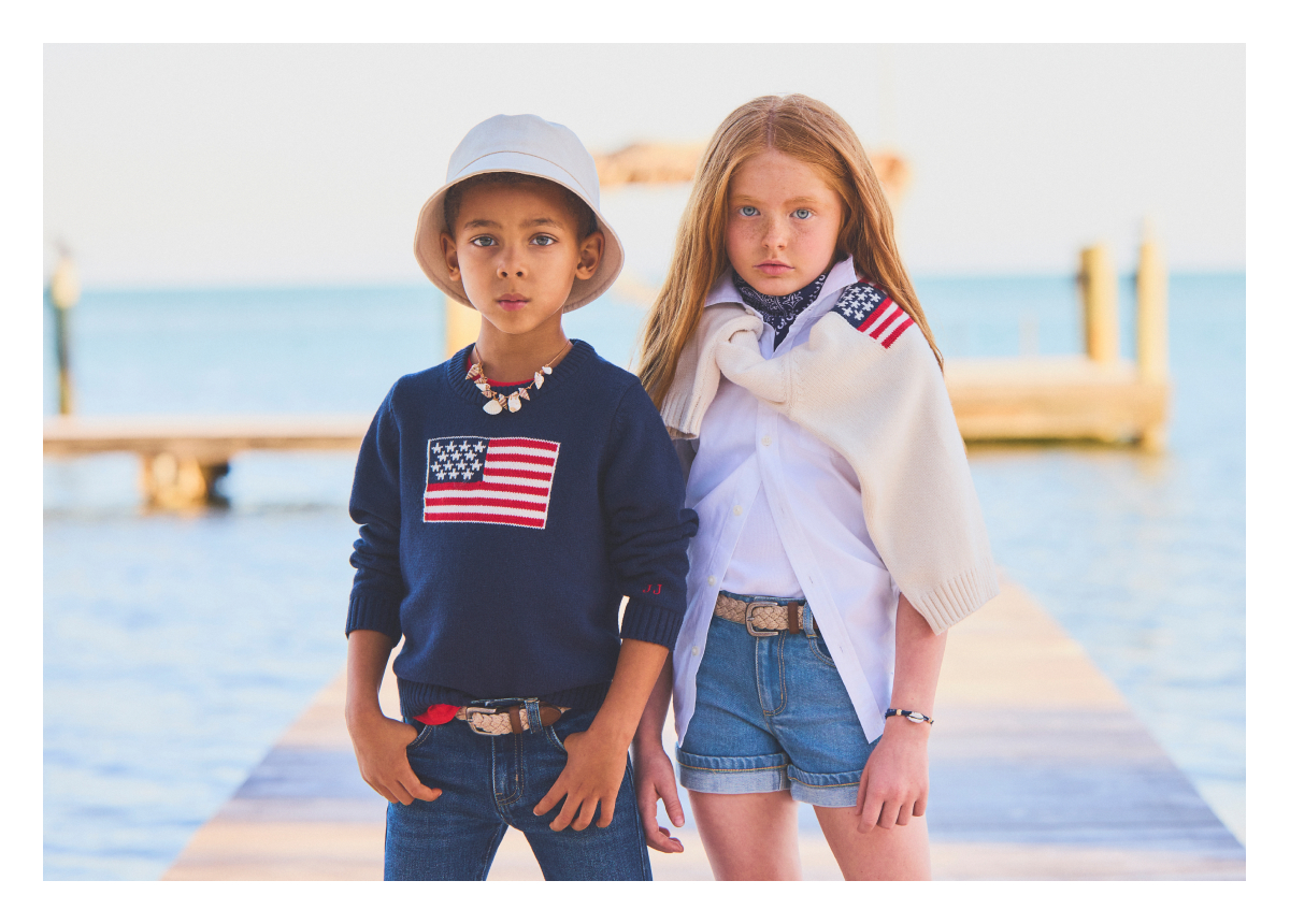 Two children wearing preppy clothing with American flag patches standing by water with boats in background