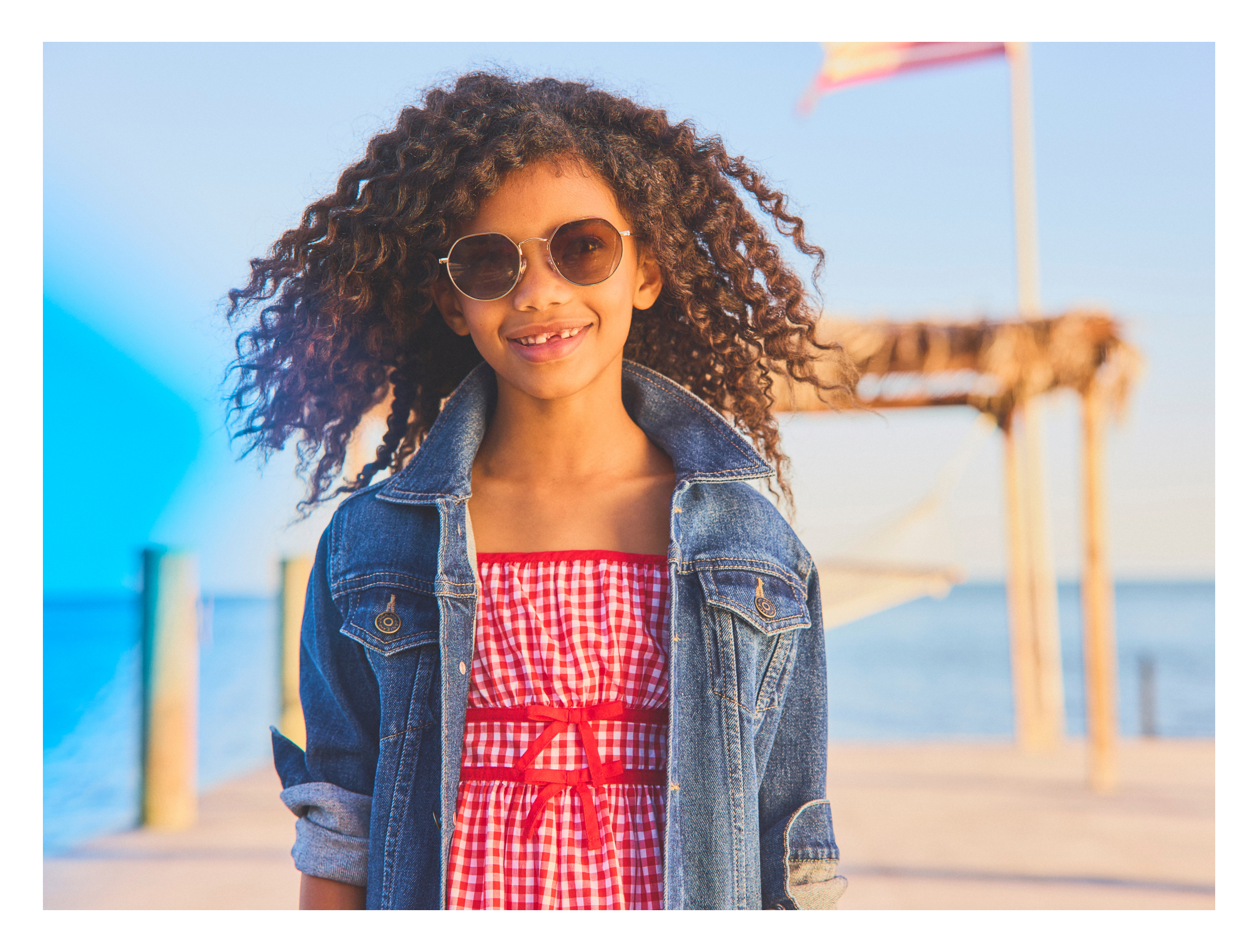 Young girl wearing sunglasses, denim jacket, and red checkered dress standing outdoors
