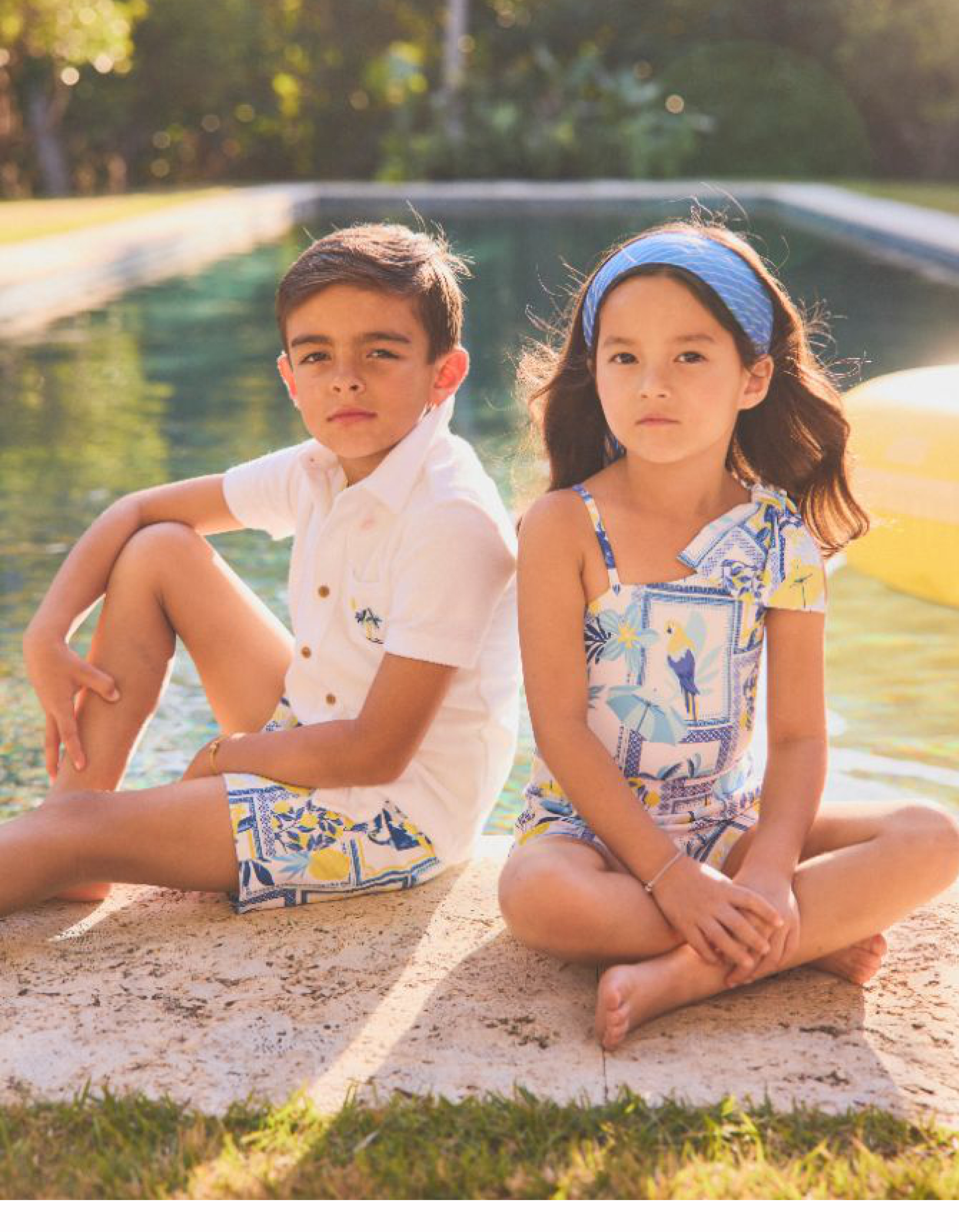 Two children sitting poolside wearing coordinating blue and yellow printed summer outfits