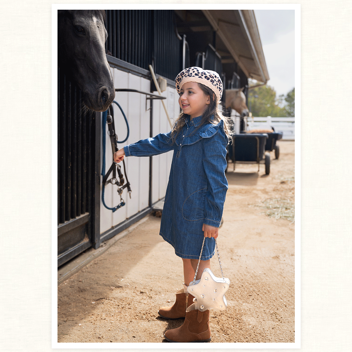 Young girl in denim dress and leopard print beret feeding a horse at a stable, carrying a star-shaped purse and wearing brown cowboy boots