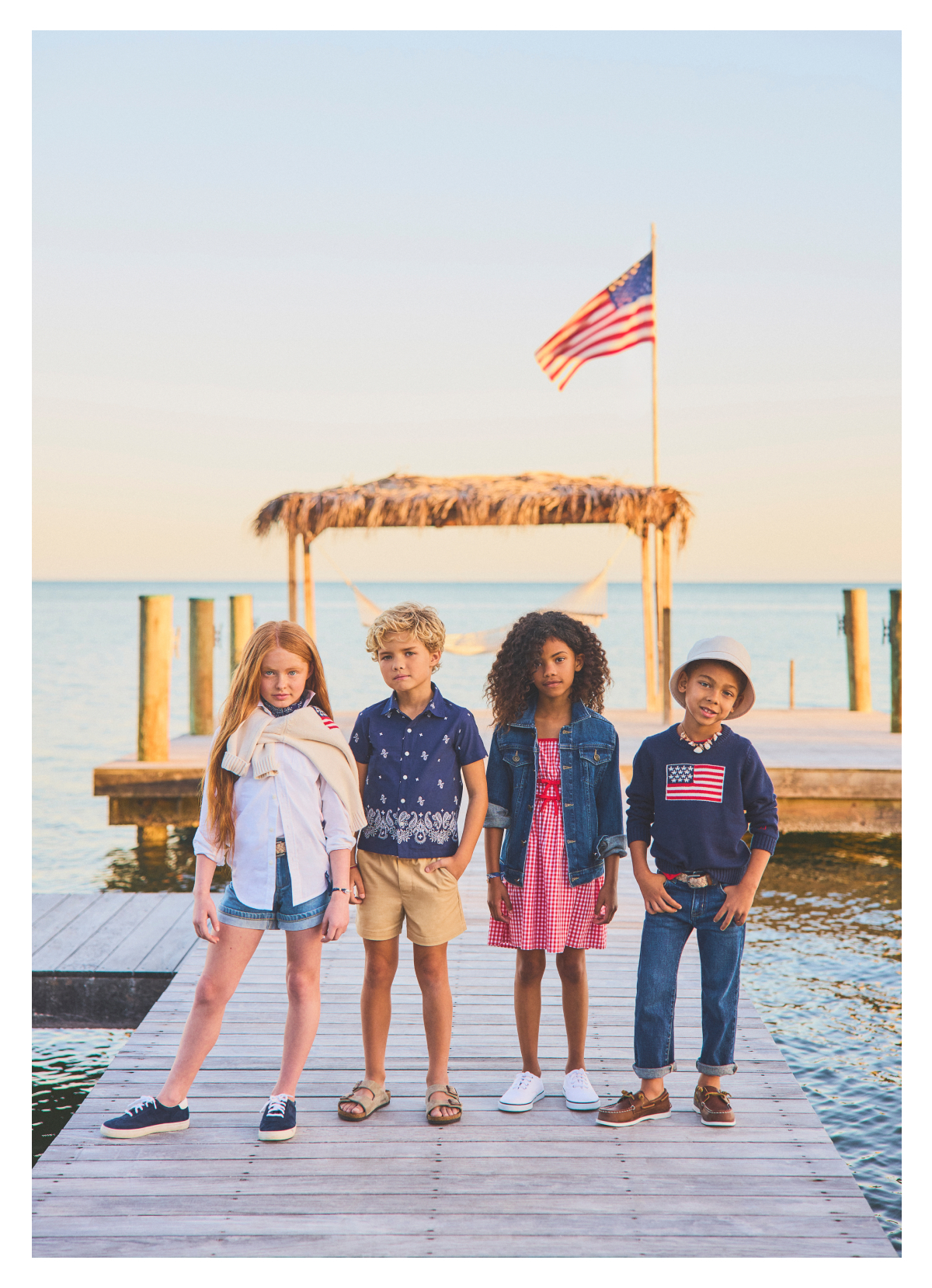 Four children wearing patriotic clothing standing on a wooden dock by water with an American flag