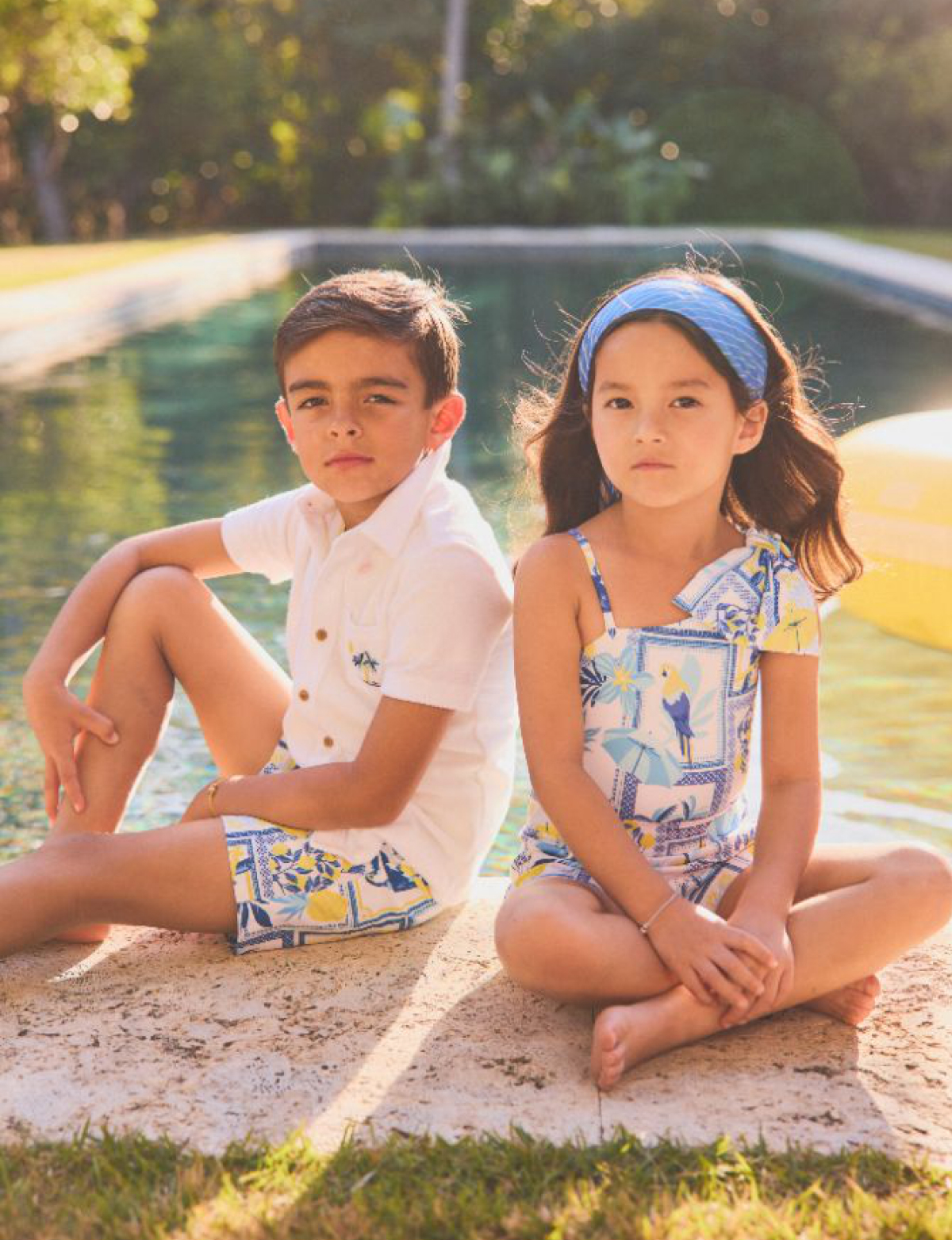 Two children sitting by a pool wearing matching blue and yellow patterned summer clothing