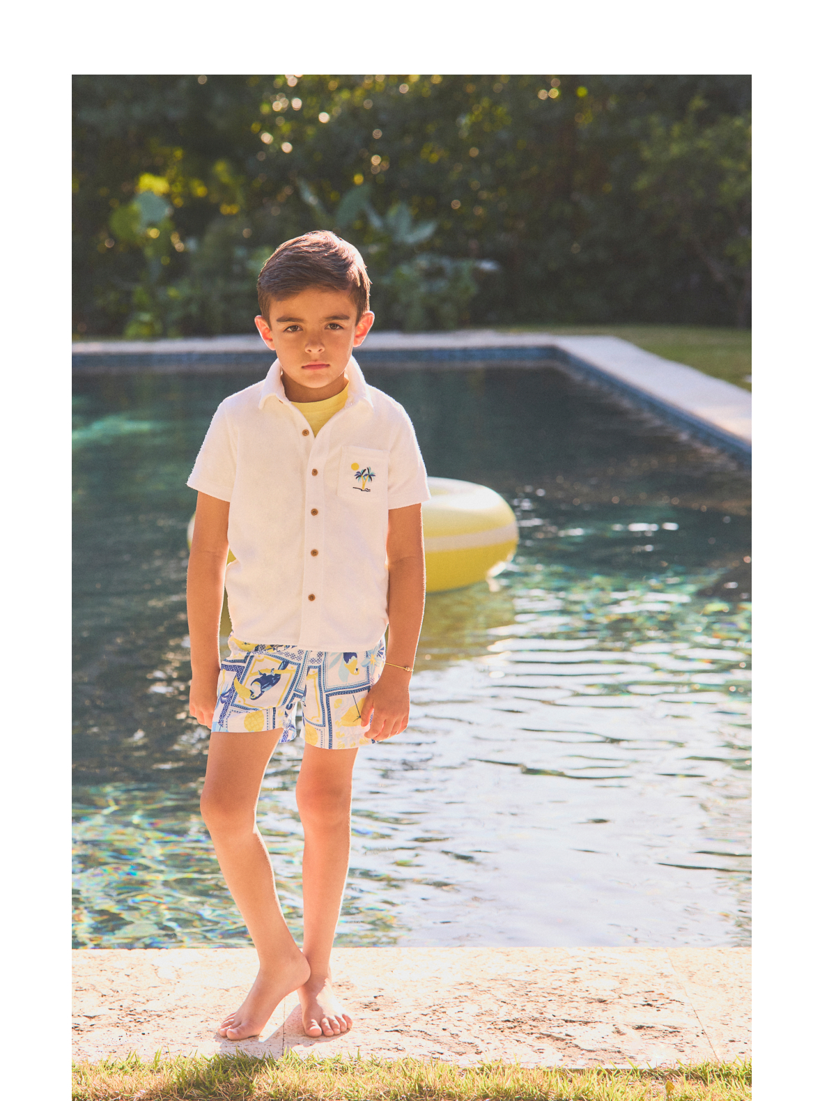 Young boy in white shirt and patterned swim shorts standing by a swimming pool