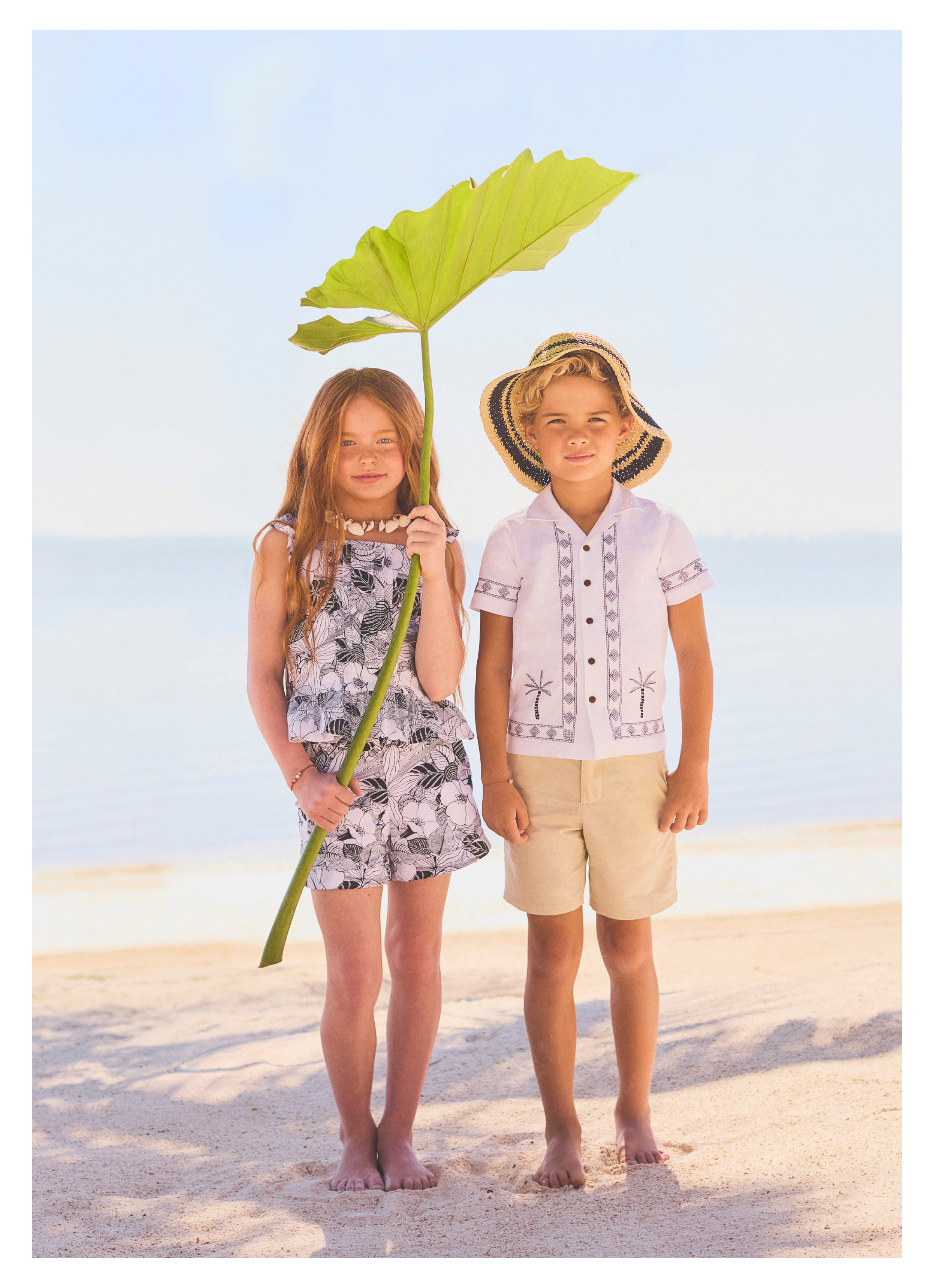 Two children standing barefoot on a beach, one holding a large tropical leaf, dressed in summer resort-style clothing