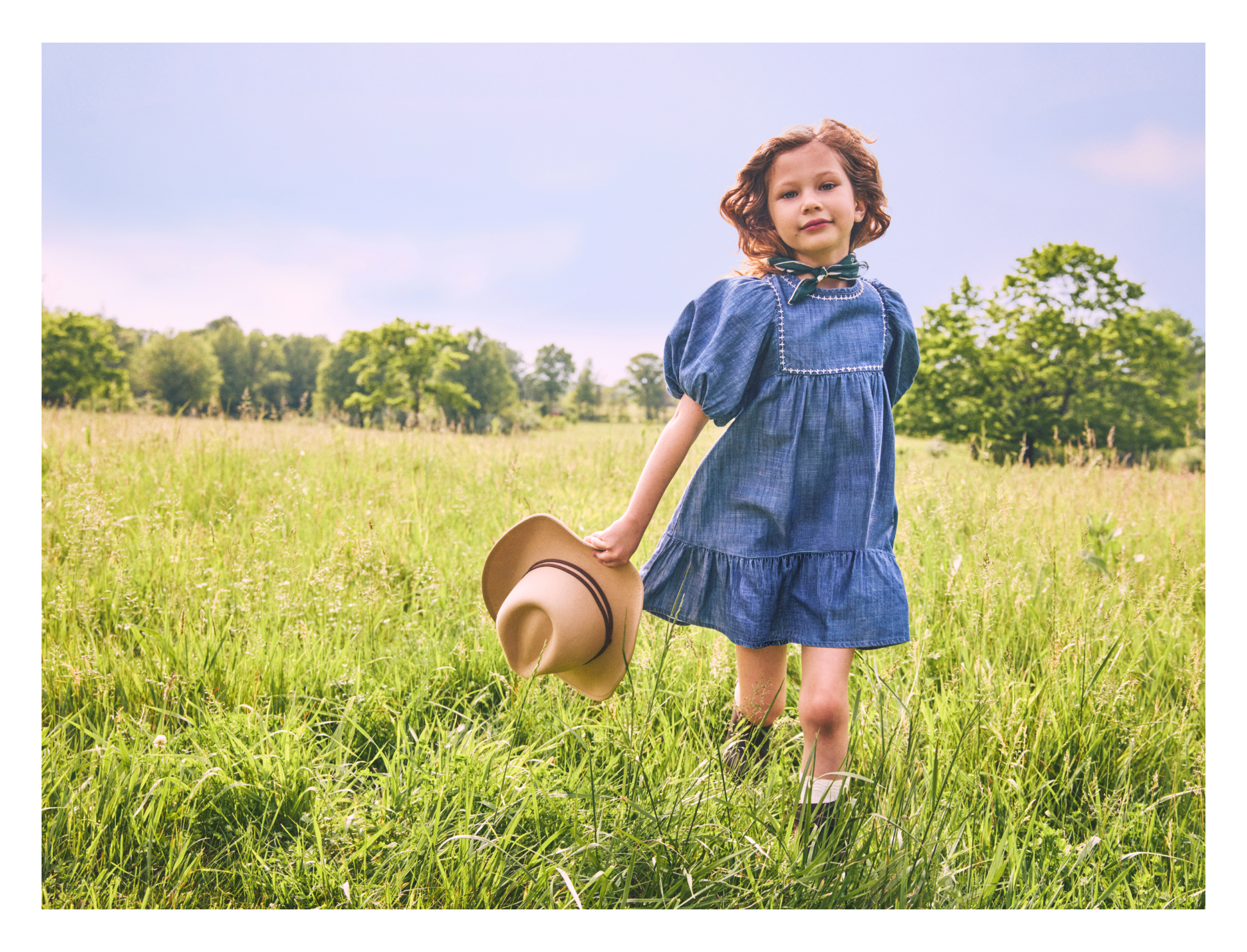 Young girl in a blue denim dress holding a straw hat while standing in a green meadow