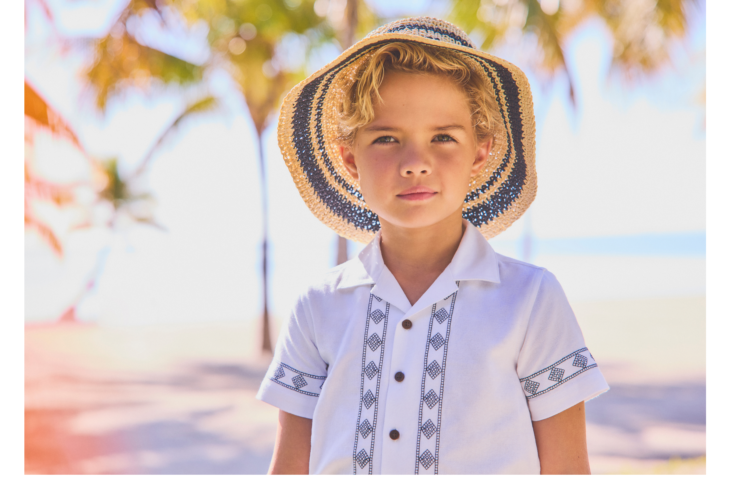 Young boy wearing a straw sun hat and white embroidered guayabera shirt, standing outdoors near palm trees