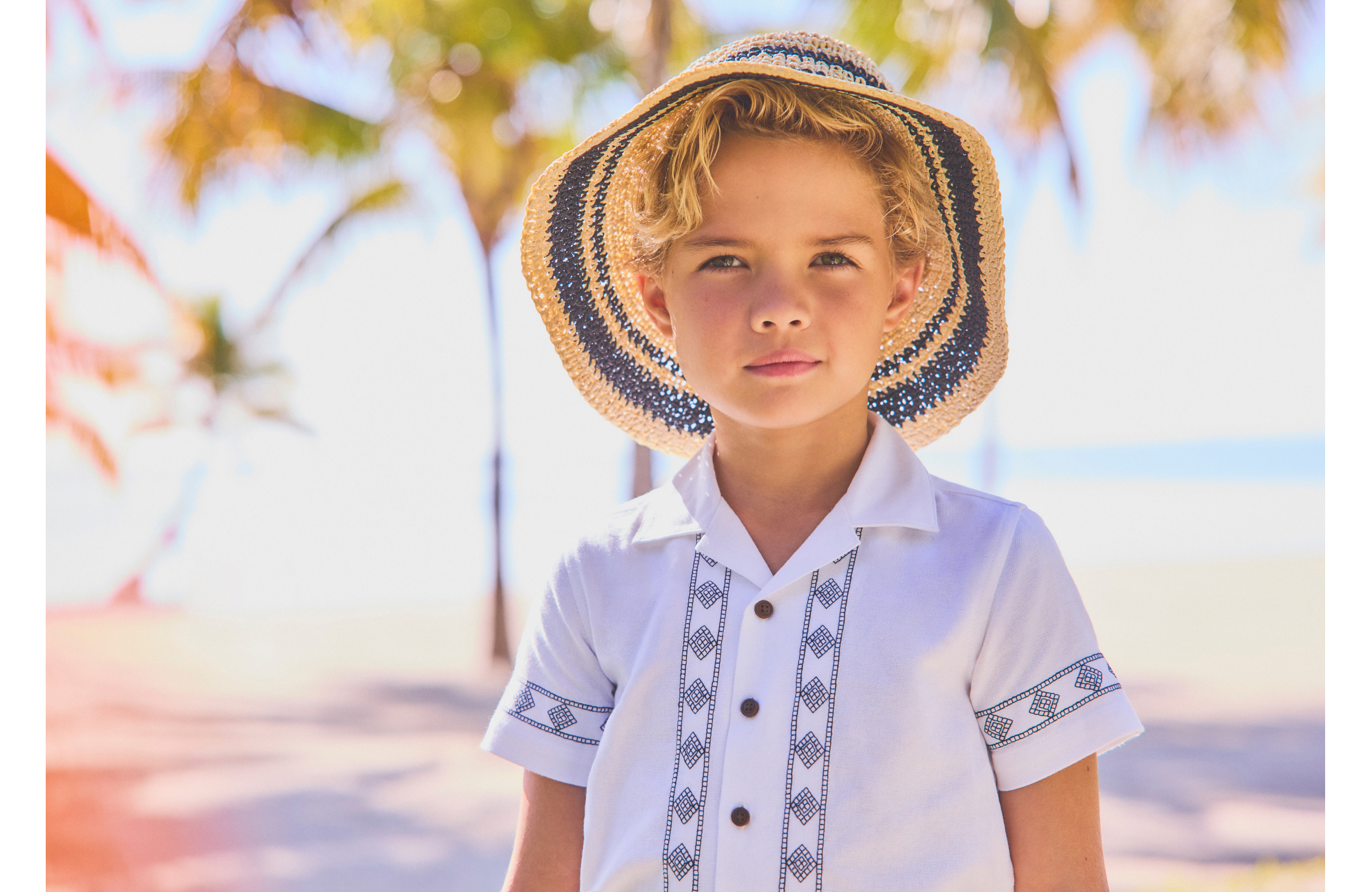 Young blonde boy wearing a striped straw hat and white embroidered guayabera shirt, standing outdoors near palm trees