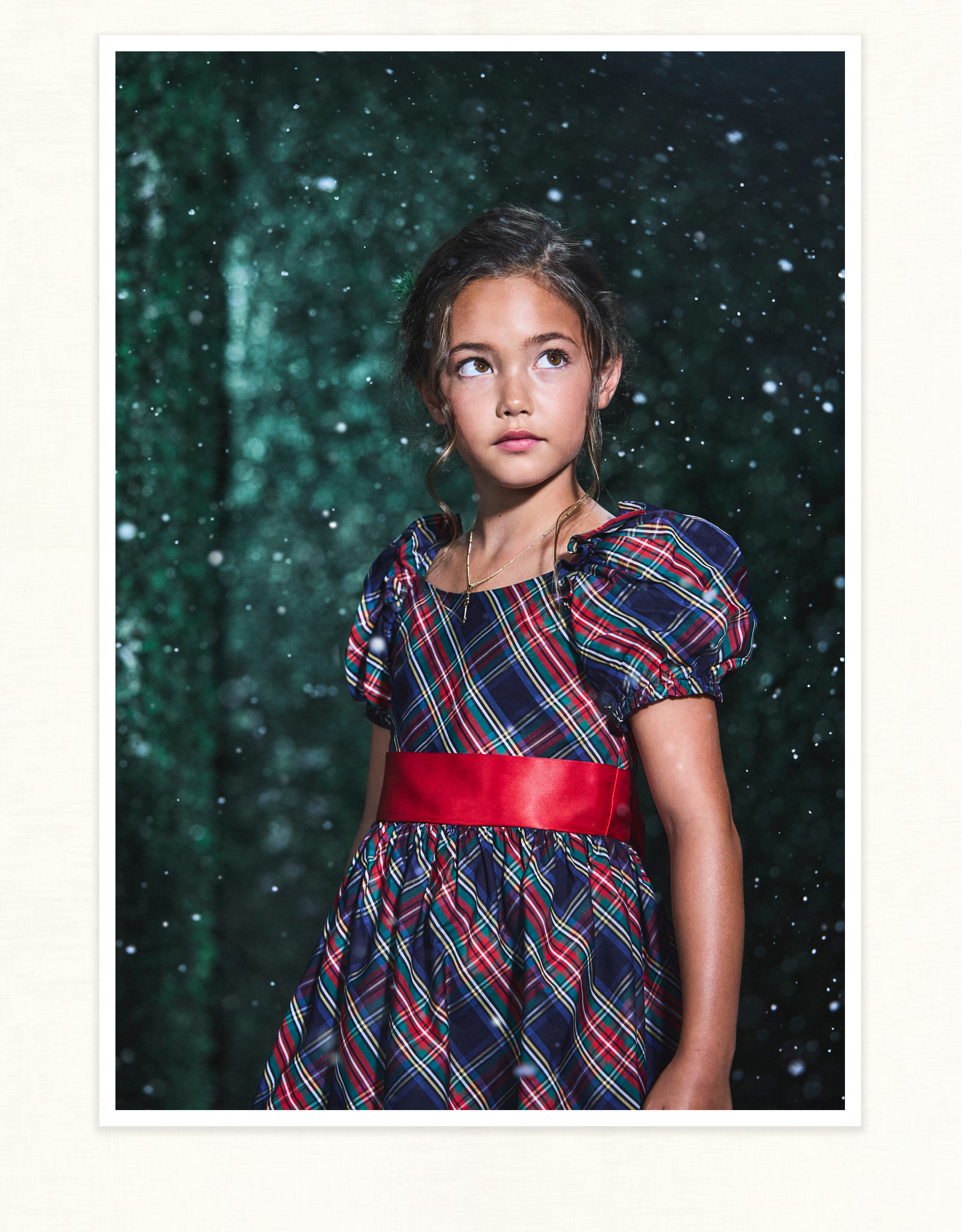 Young girl in a tartan plaid dress with red sash, looking upward, with a snowy green background