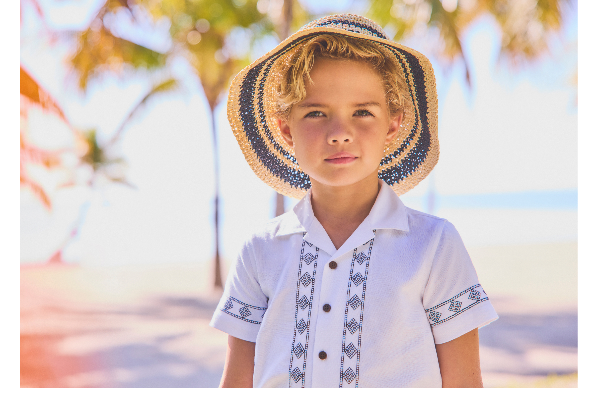 Young blonde boy wearing a striped straw sun hat and white embroidered guayabera shirt, standing outdoors near palm trees