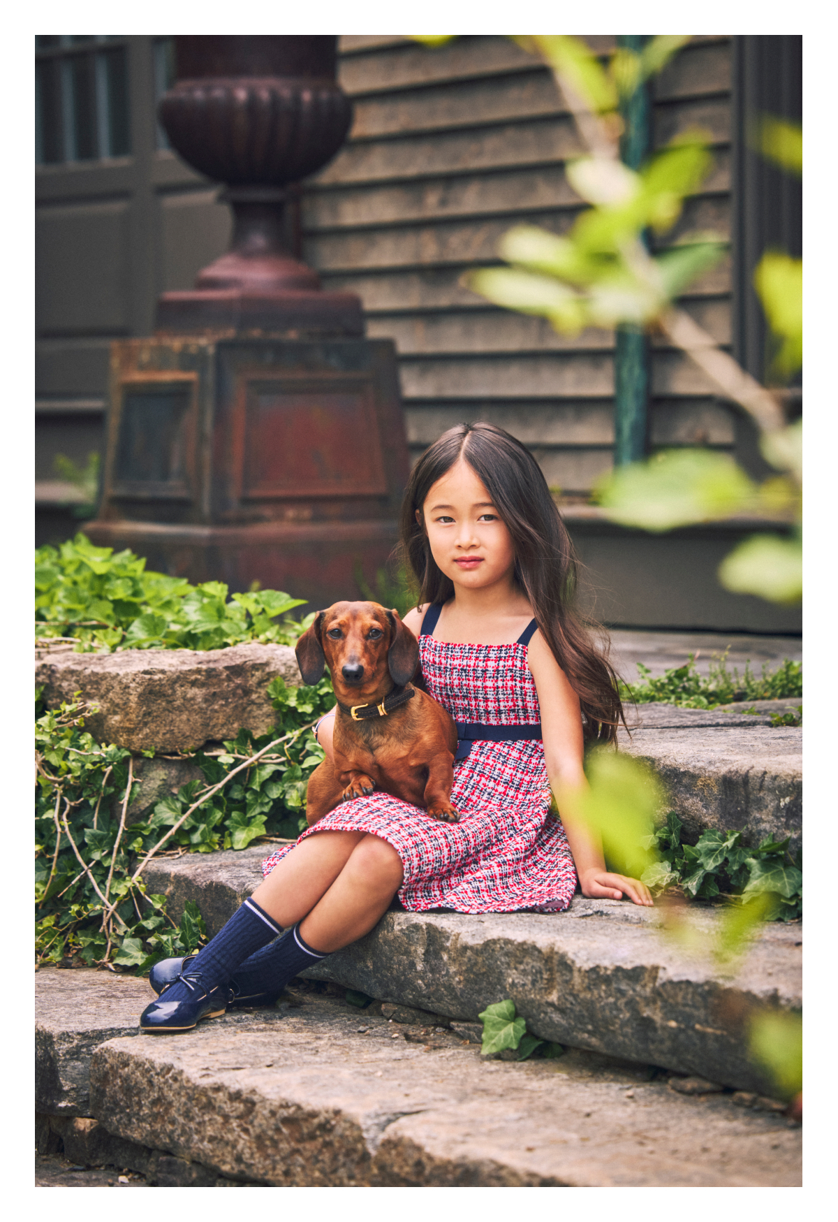 A young girl in a red tweed dress sitting on stone steps holding a dachshund puppy