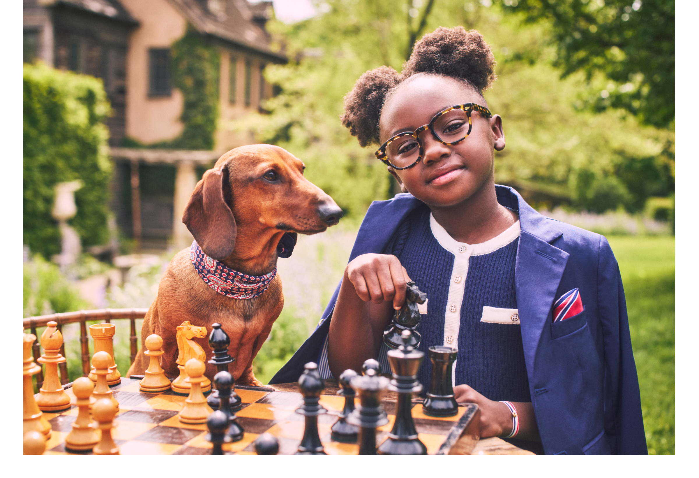 Young girl in navy blazer playing chess outdoors with a dachshund wearing a bandana