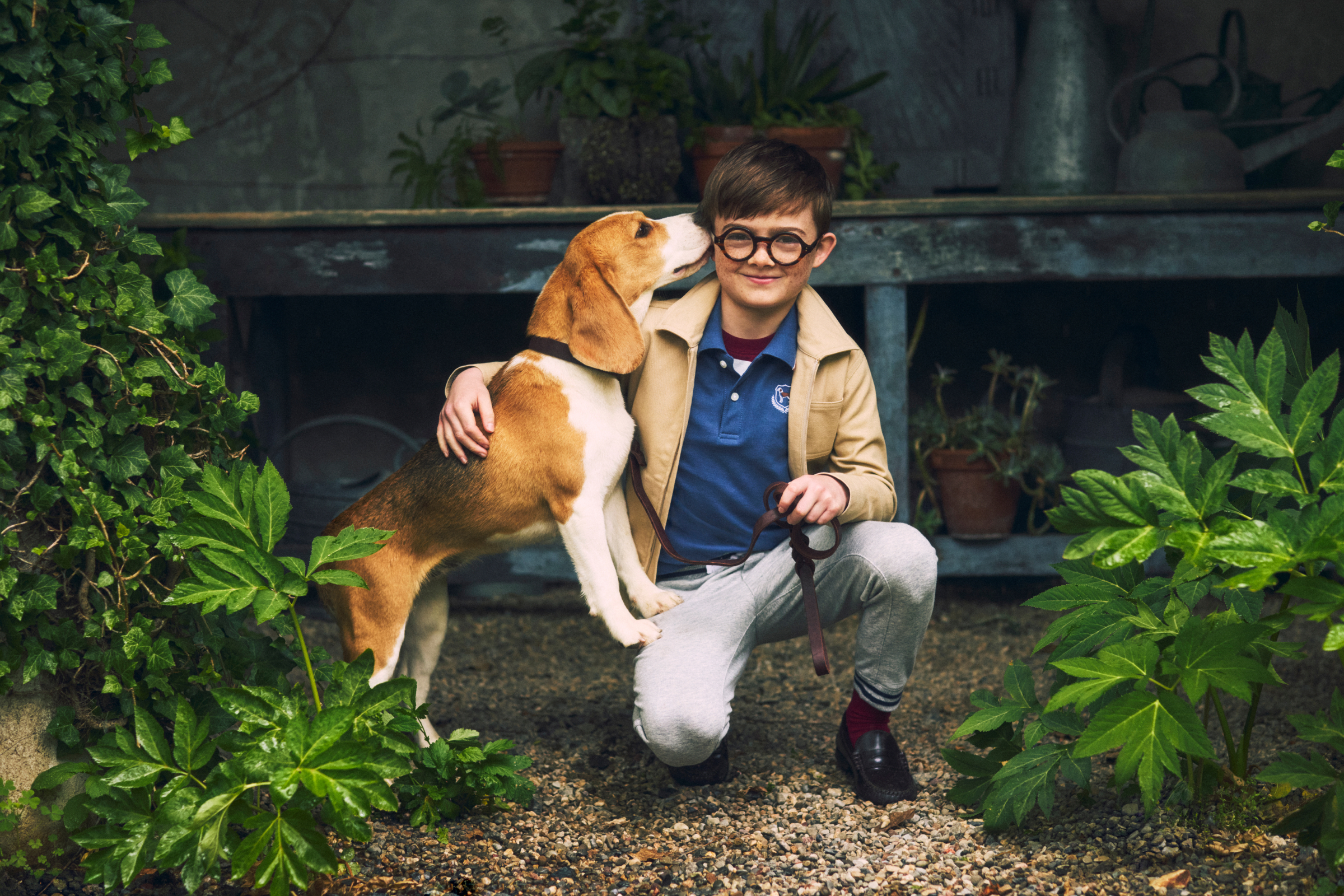 A boy with round glasses crouching outdoors with a beagle puppy nuzzling his face, surrounded by green plants