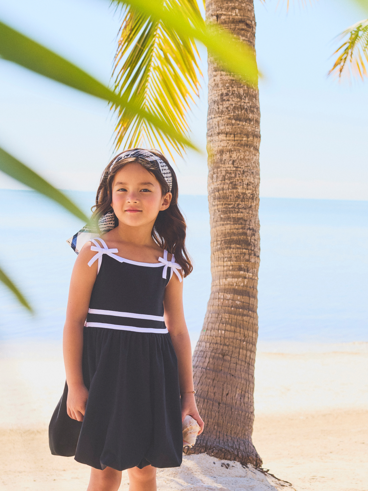Young girl in black dress with white trim standing by palm tree on beach