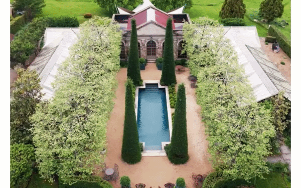 Aerial view of an estate with a rectangular pool, tall cypress trees, and a stone building with a red roof surrounded by lush greenery