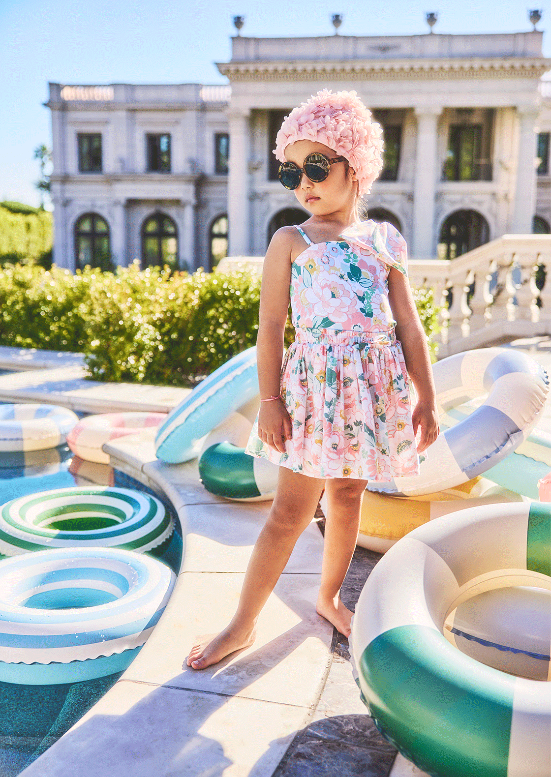 Young girl in floral dress and pink swim cap wearing sunglasses standing among colorful pool floats