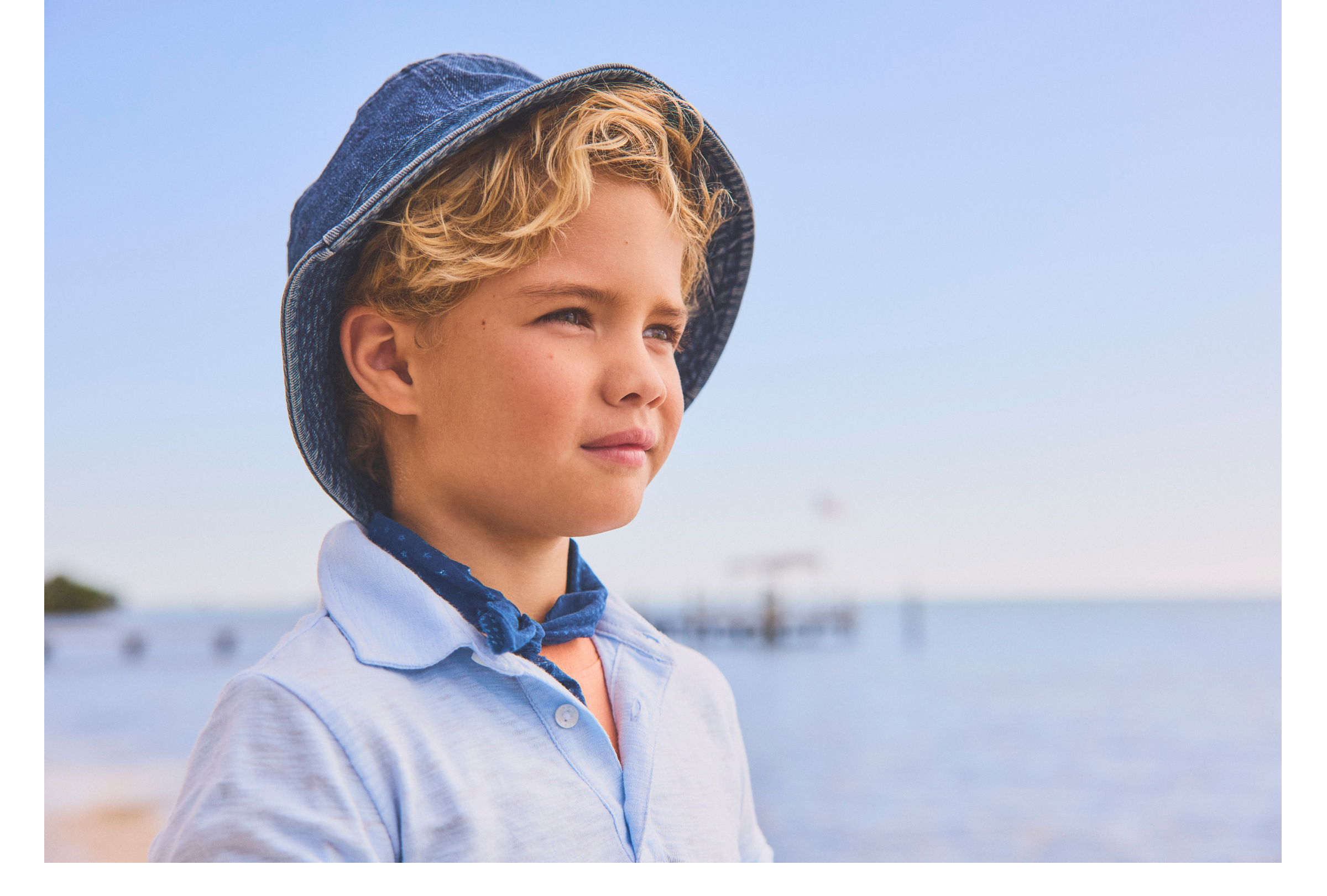 Young blond boy wearing a denim bucket hat and light blue shirt with a navy bandana, gazing into the distance at the beach