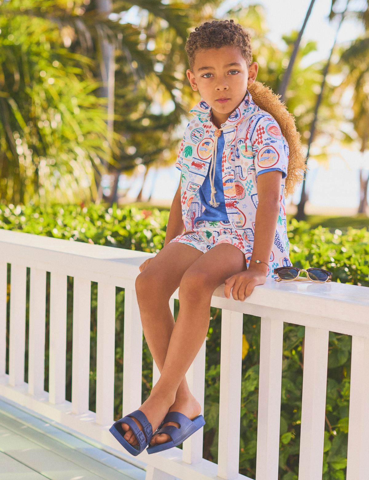 Young boy in colorful patterned shirt and blue sandals sitting on white railing outdoors