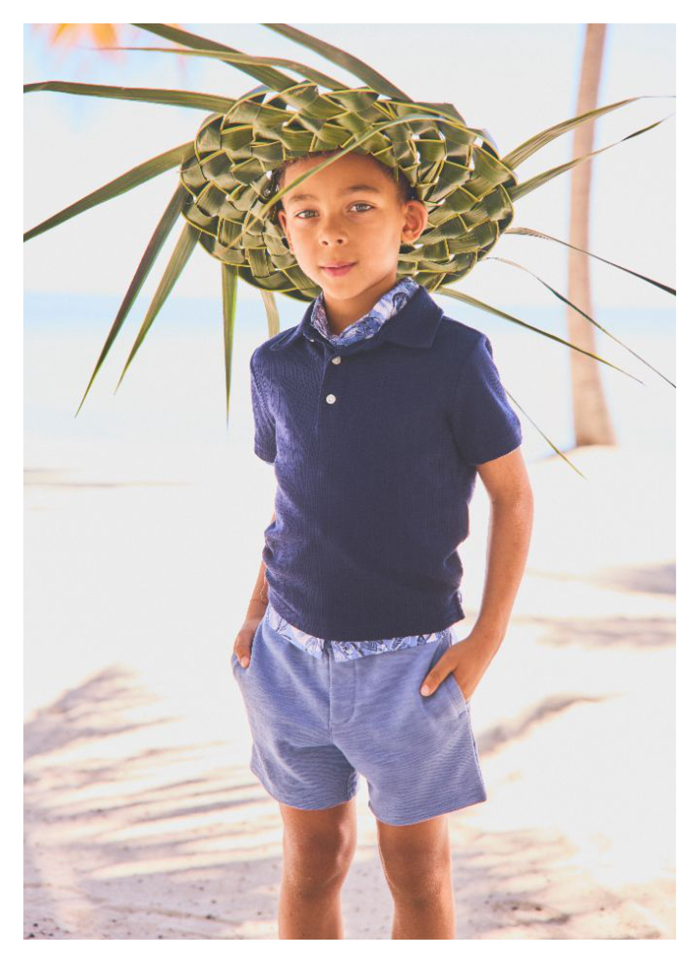 A young boy wearing a woven palm leaf hat, navy polo shirt, and blue shorts standing on a beach