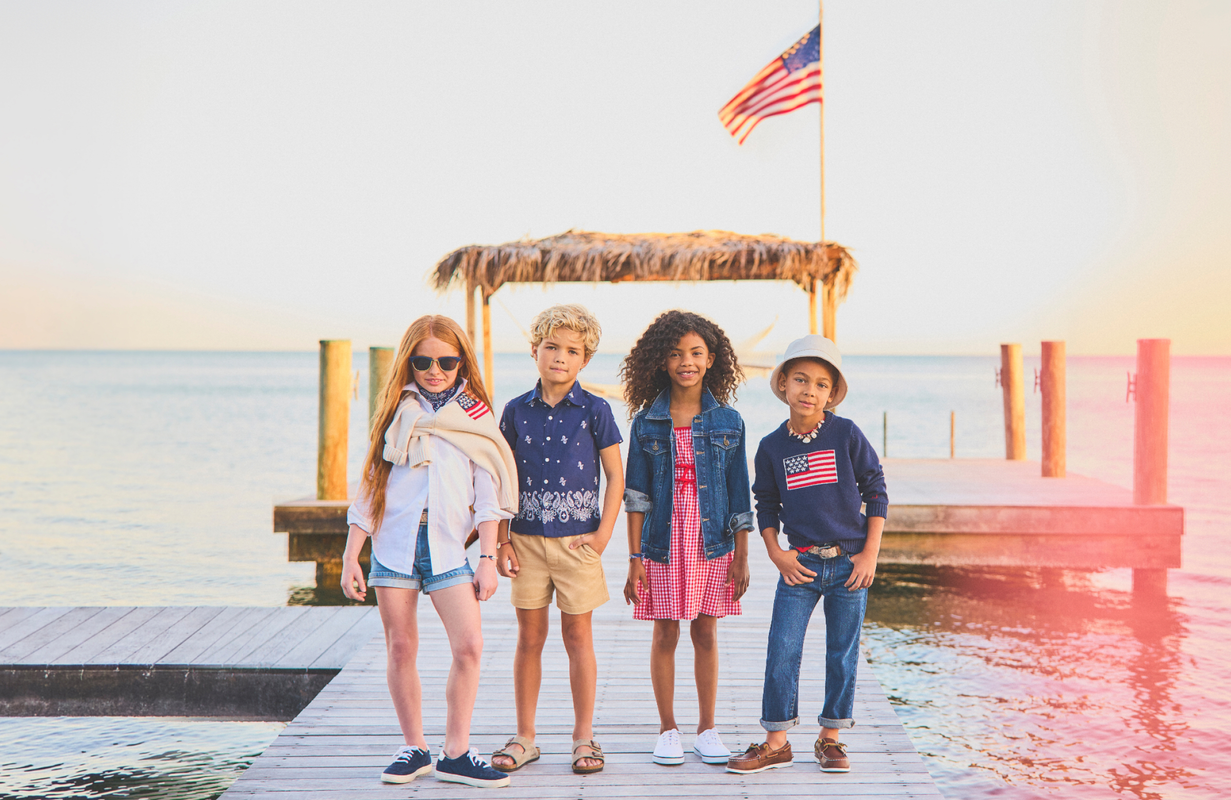 Four children posing on a waterfront dock at sunset, wearing American flag-themed summer outfits, with a US flag and thatched gazebo in the background