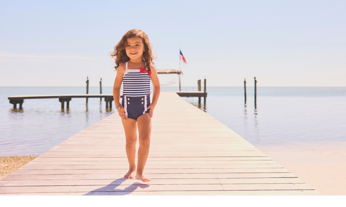 Young girl in a navy striped one-piece swimsuit standing on a wooden dock by the water on a sunny day