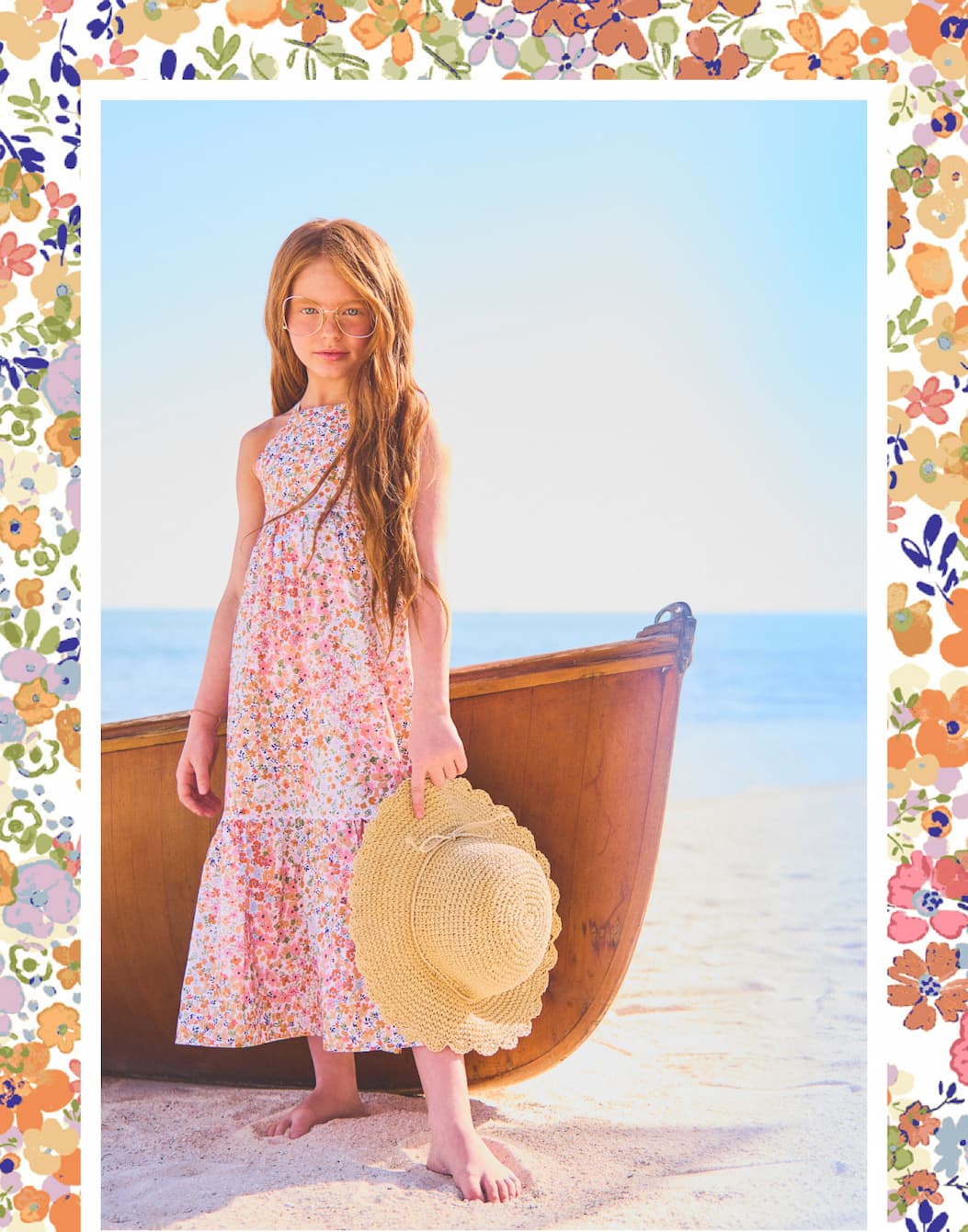Young girl in floral dress holding sun hat standing next to wooden boat on beach