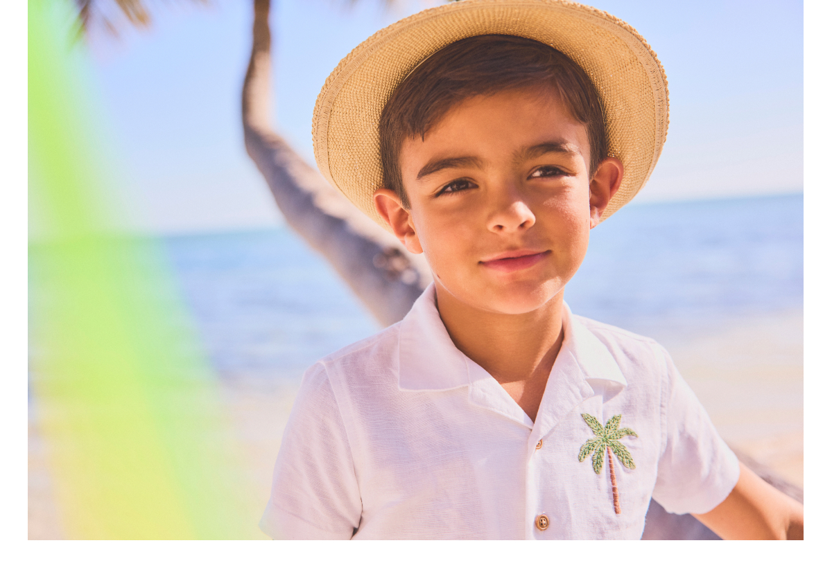 A young boy wearing a straw hat and white shirt with a palm tree embroidery, smiling at the beach
