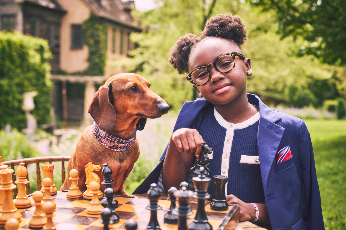 A young girl wearing glasses and a navy blazer playing chess outdoors with a dachshund wearing a bandana