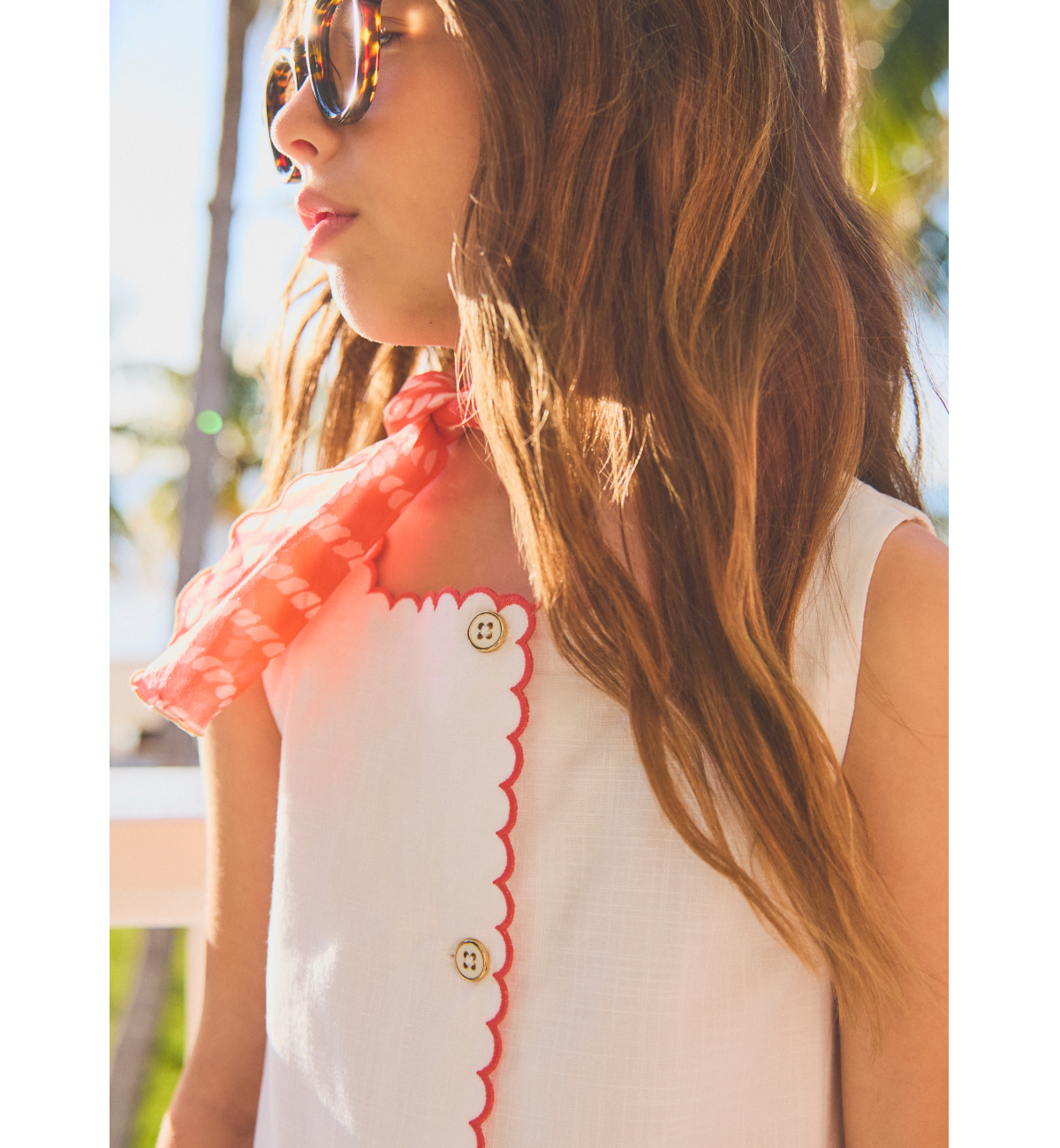 Woman in white scallop-trim button-front dress with coral neck scarf and tortoiseshell sunglasses, outdoors in sunny setting