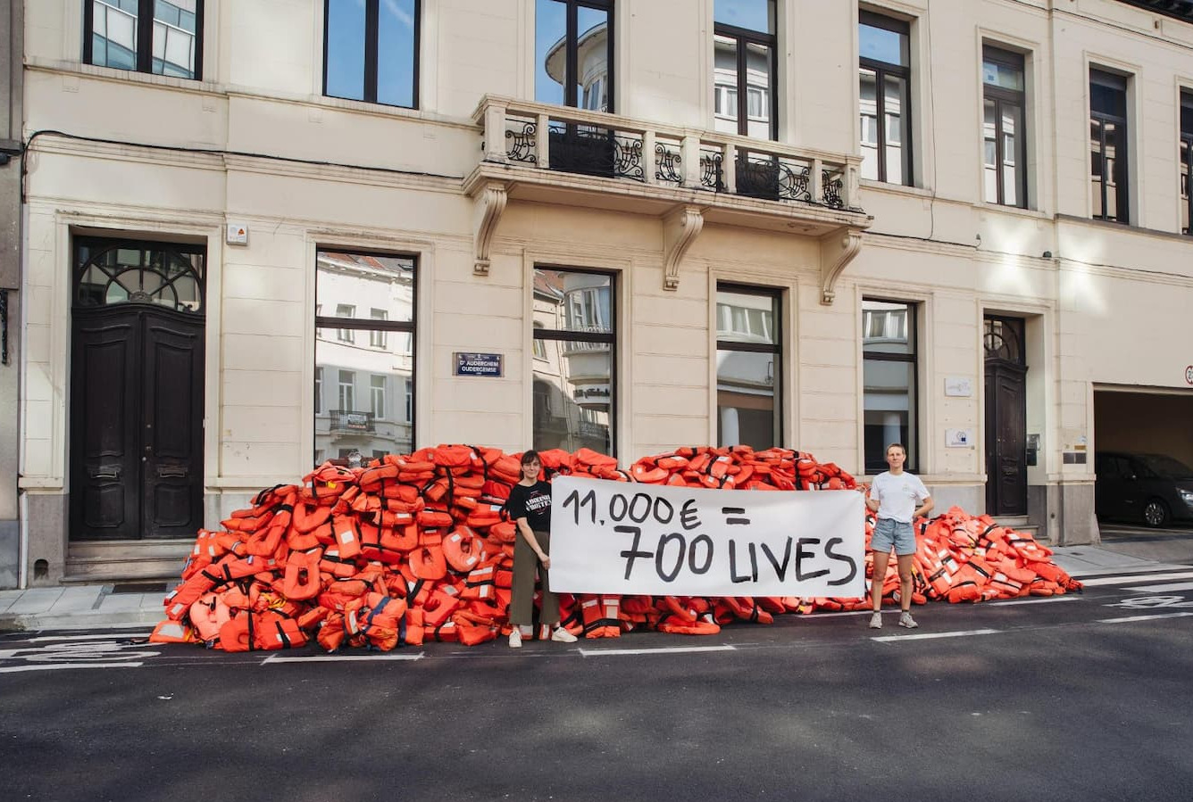 700 Rettungswesten liegen vor einem Bürogebäude, davor zwei Personen mit einem Banner auf dem steht: 11.000 € = 700 Lives